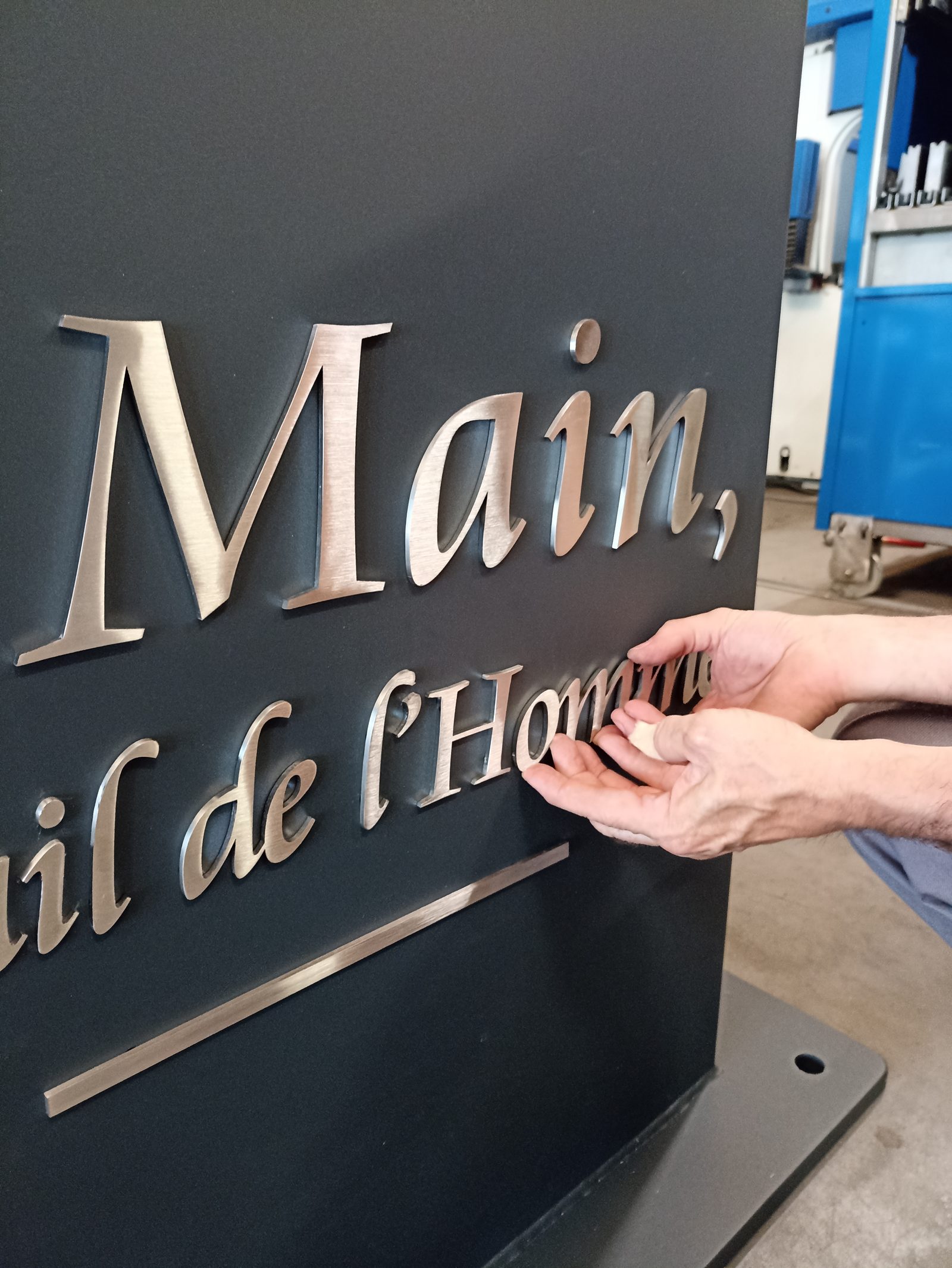 Les mains d'un compagnon ajustent des lettres d'inox brossé sur une stèle anthracite